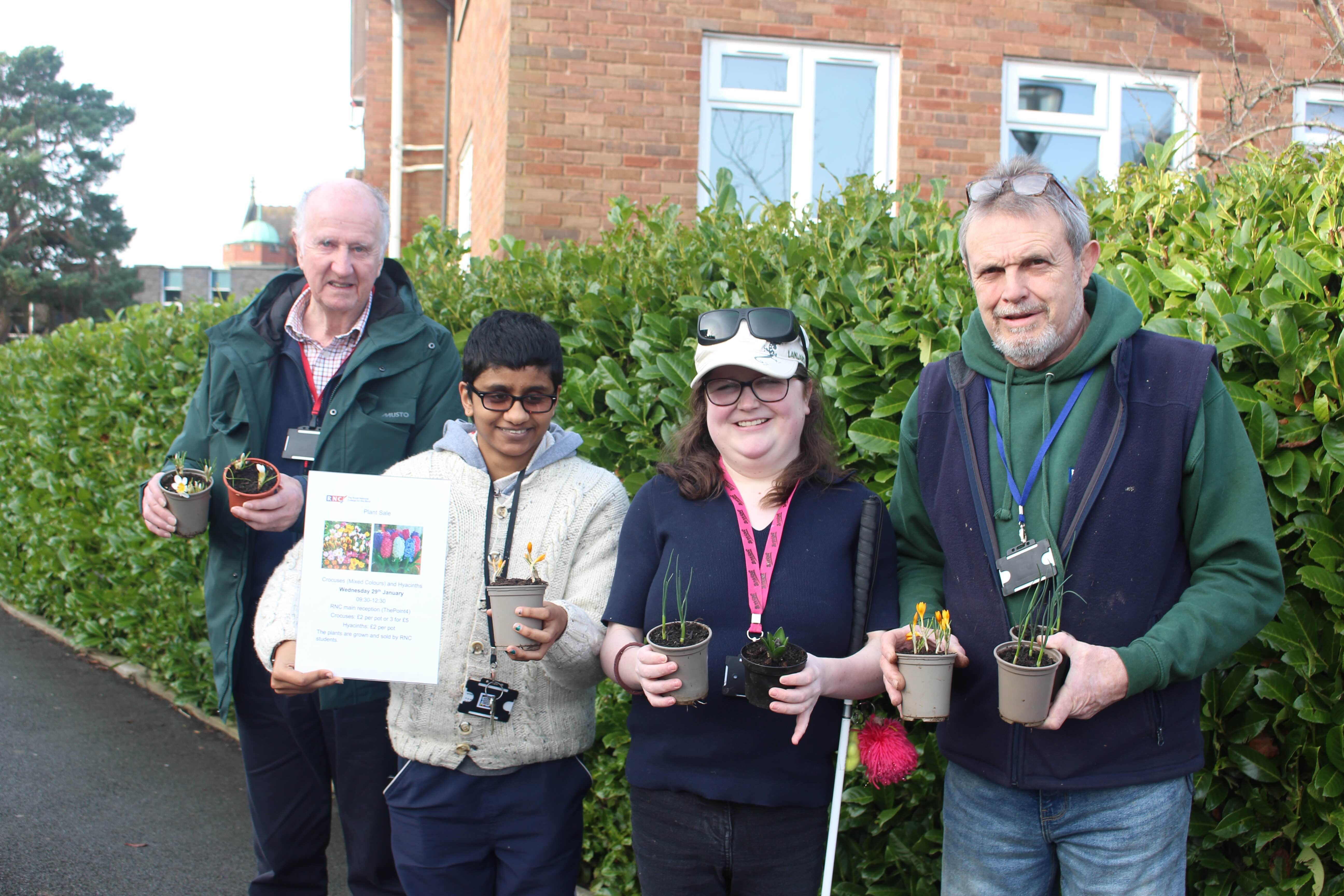 gardening volunteers and students standing by a laurel hedge holding plants and a poster advertising the plant sale