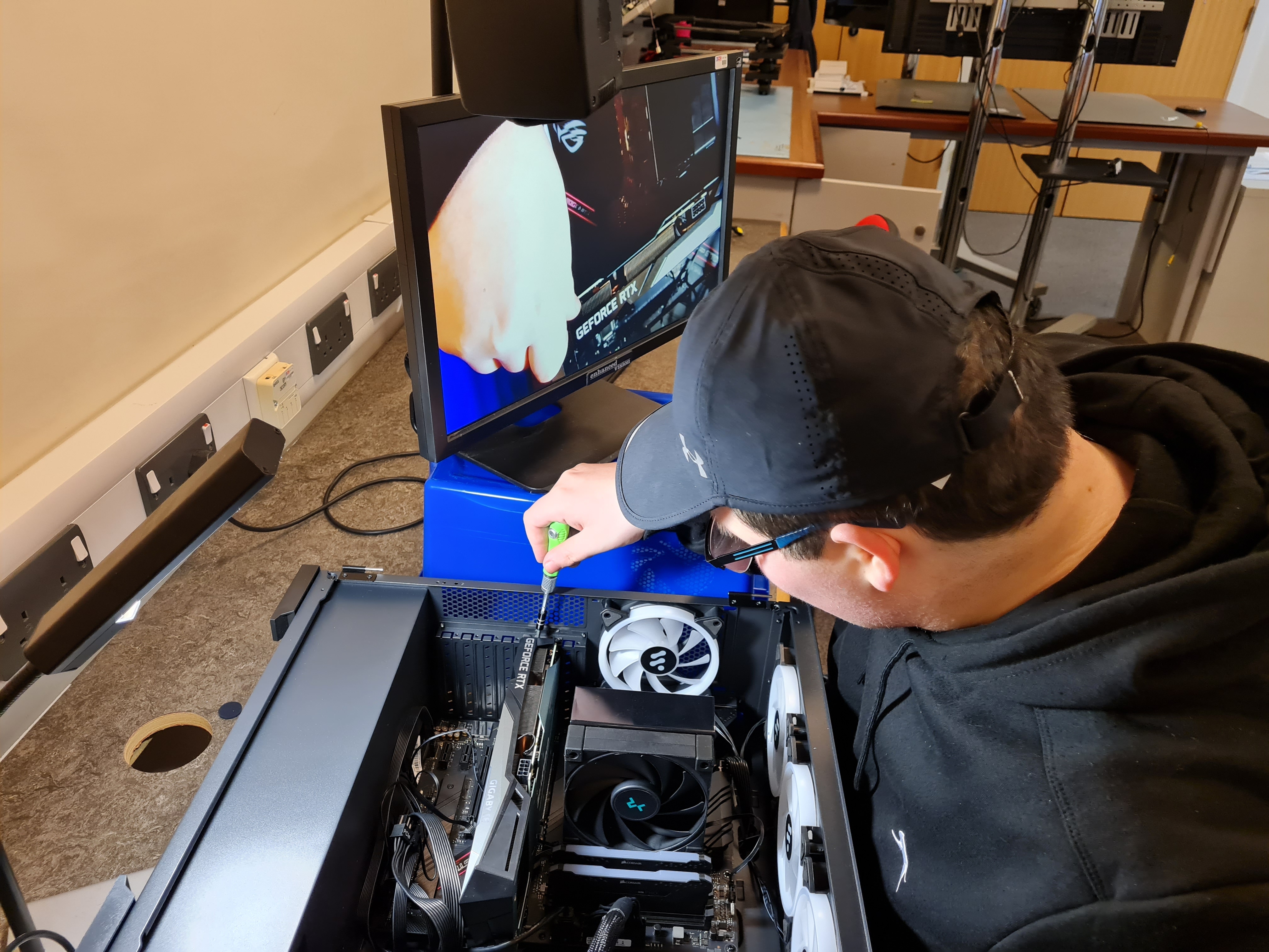 A student works on the inside of a computer box with a screwdriver - there is a magnifying monitor next to him on the right