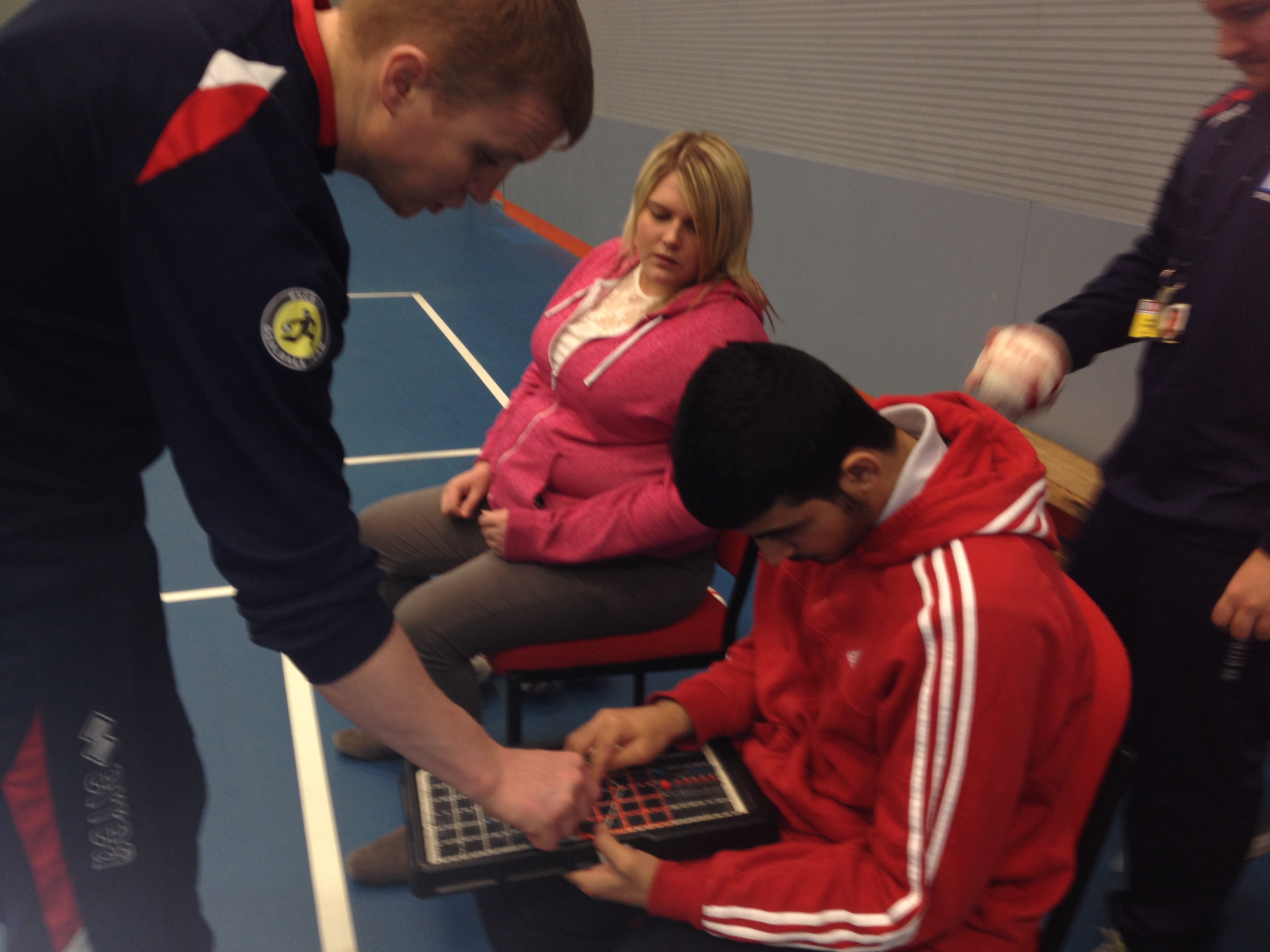 a male boccia player is plotting on the tactile map, a male teacher helps and a female player looks on 