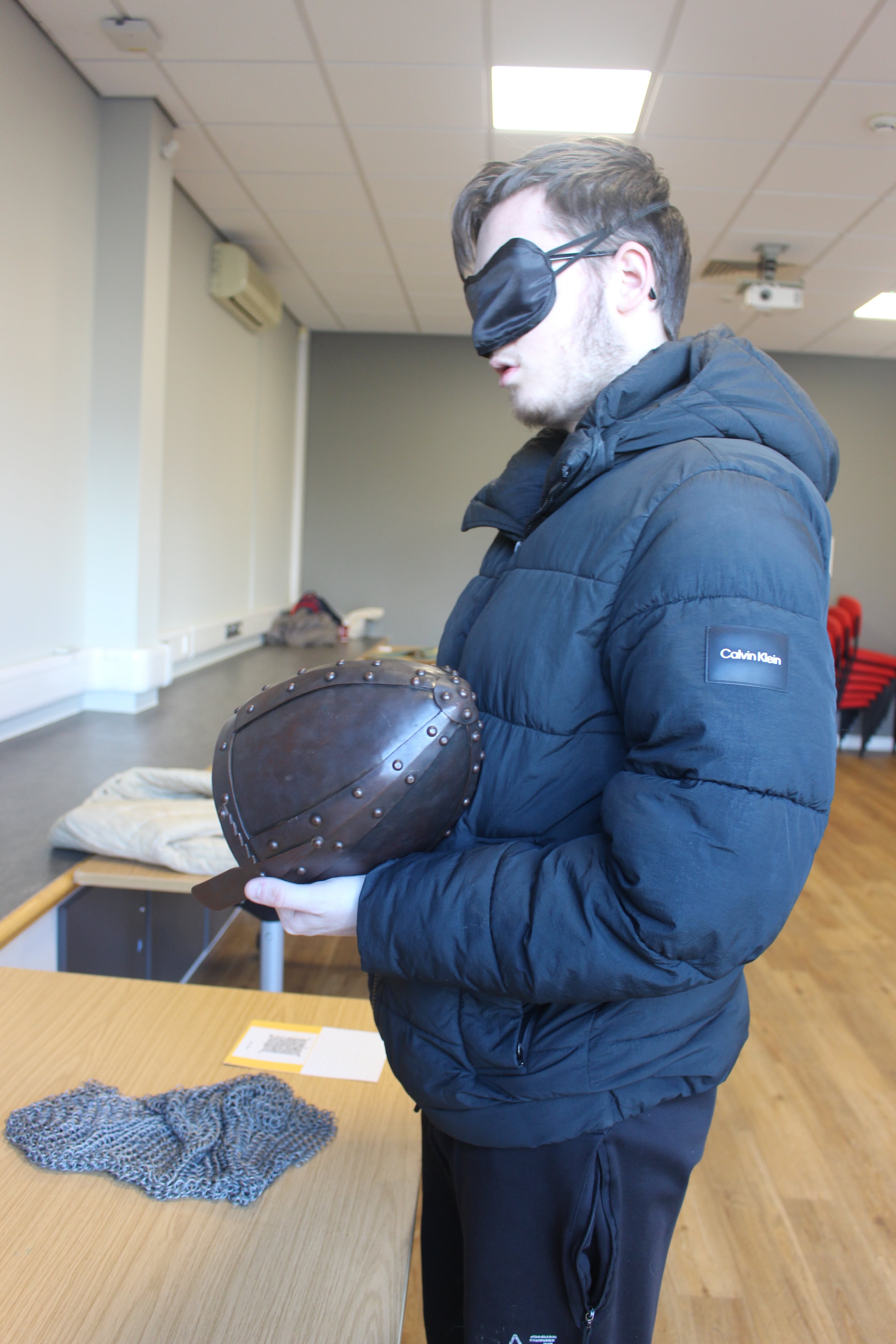 a blindfolded visitor handling a medieval helmet which has rivets and a nasal plate