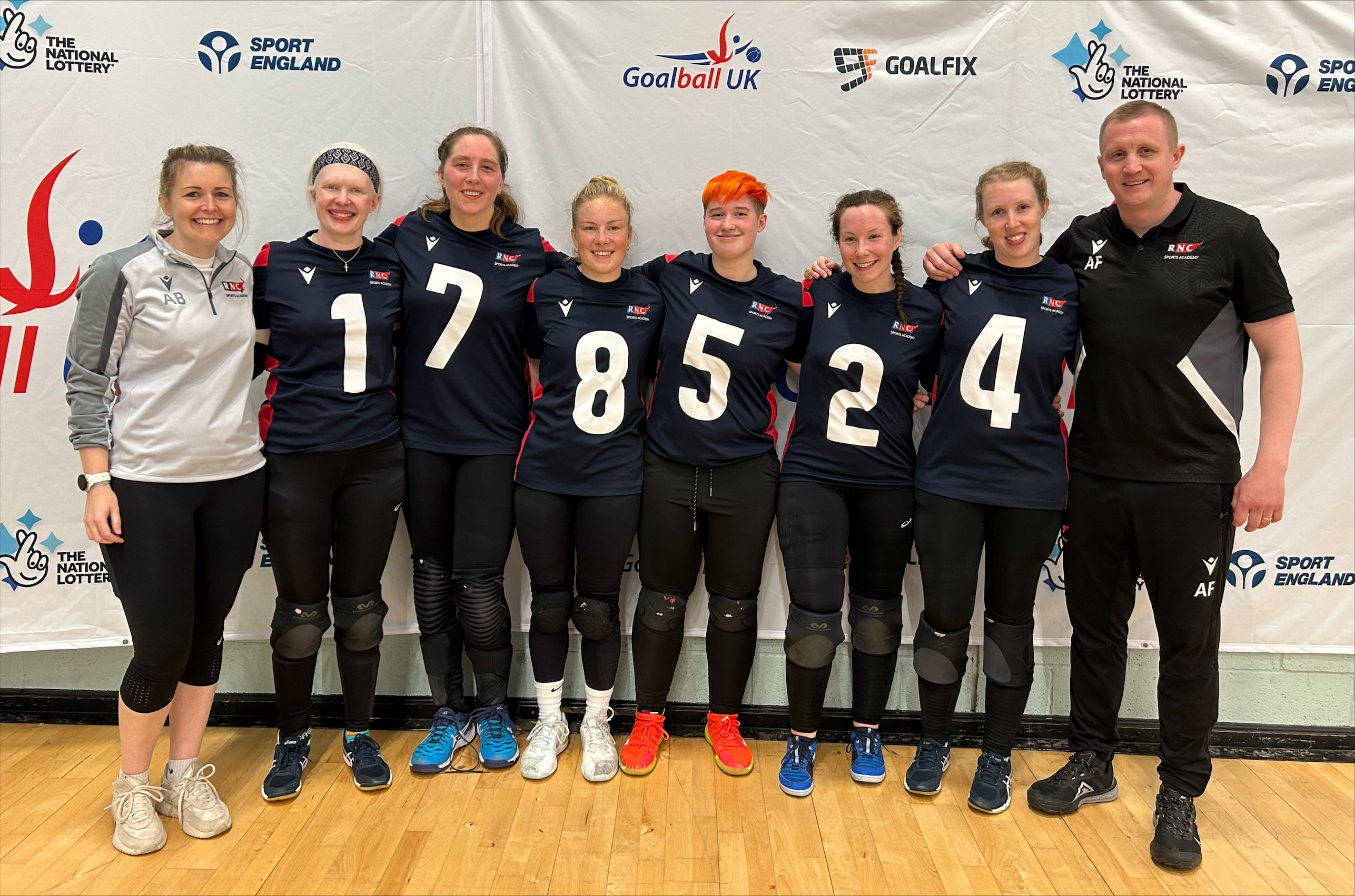 the team and coaches standing in a row in front of a sponsorship board at the event