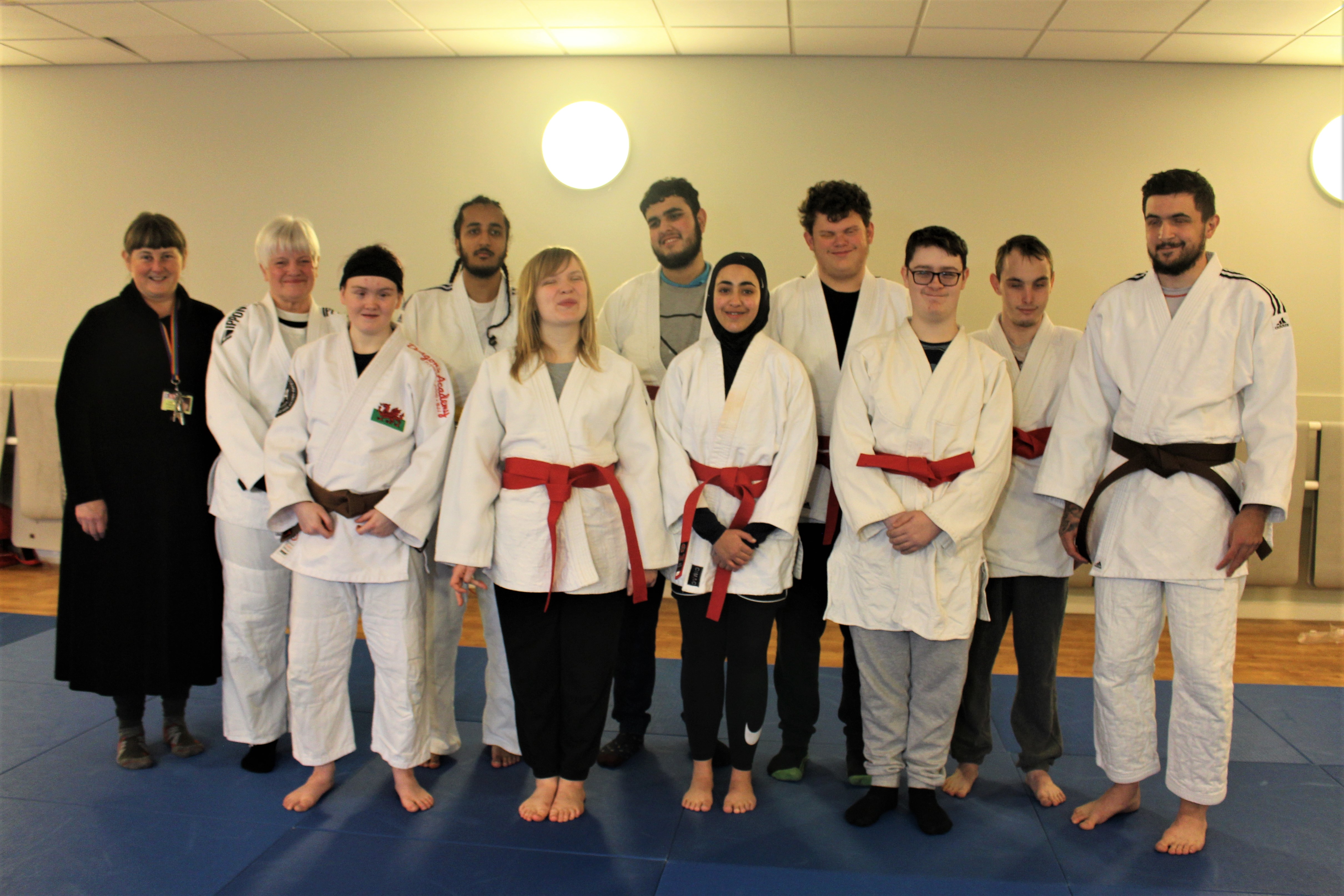 A group of students with Coach and Principal standing barefoot on Judo mats displayng their newly awarded Judo belts