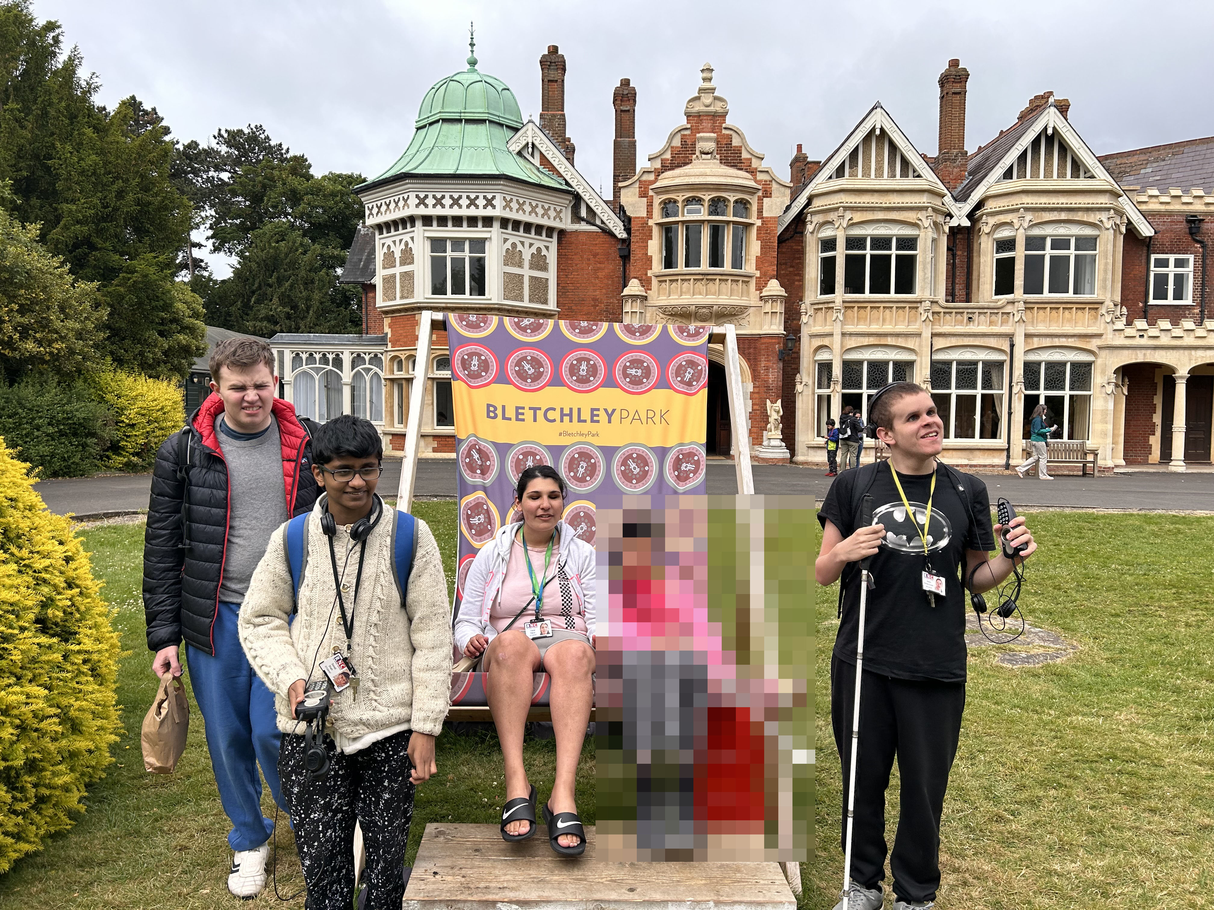 3 students stand and 2 students sit (one blurred out) on a giant deckchair in front of Bletchley Manor House 