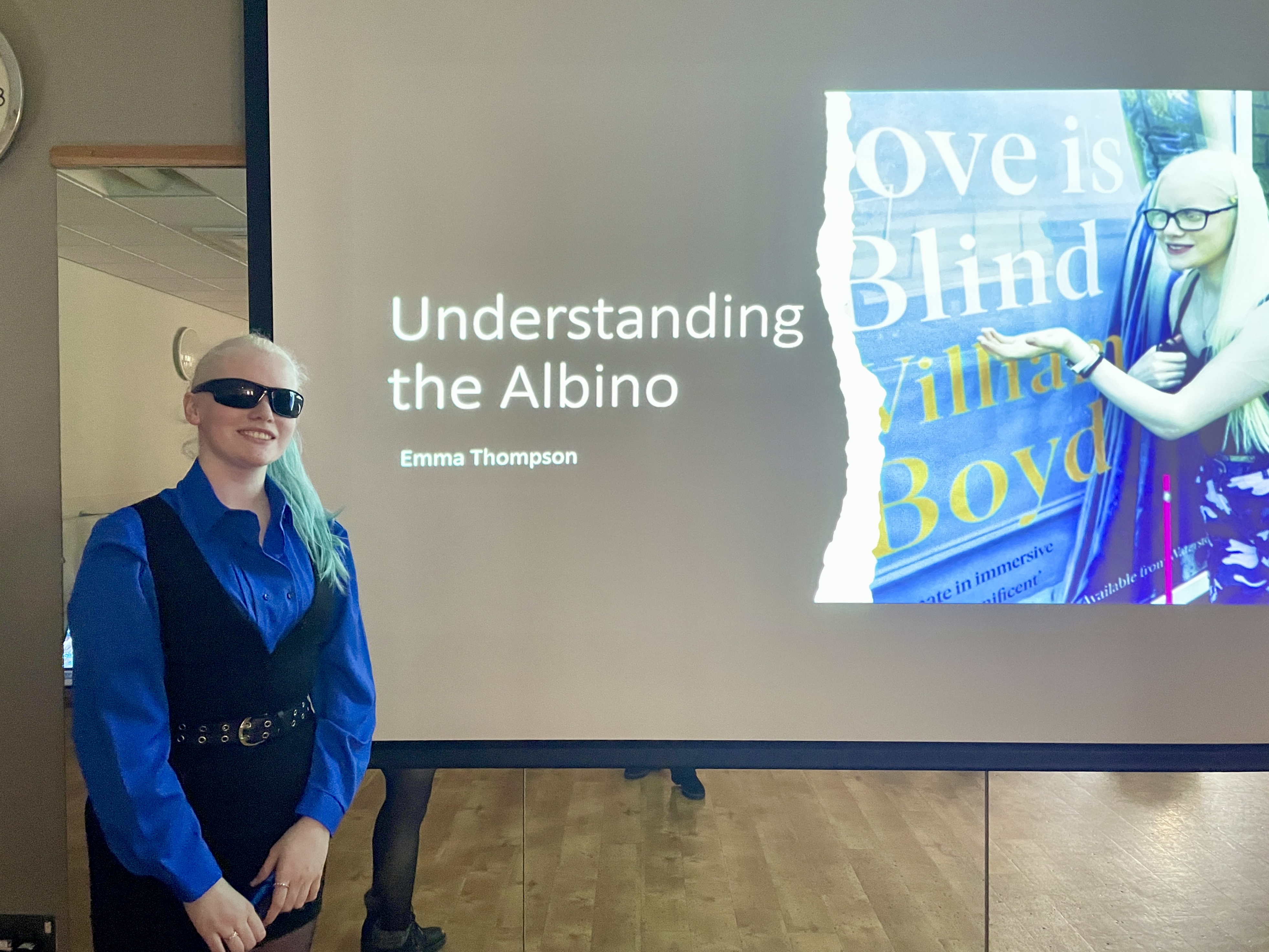 Emma stands in front of her presentation on a large screen - the slide reads: Understanding the Albino