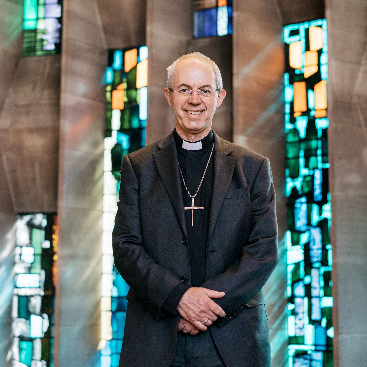 The Archbishop stands smiling in front a beautiful coloured stained glass window