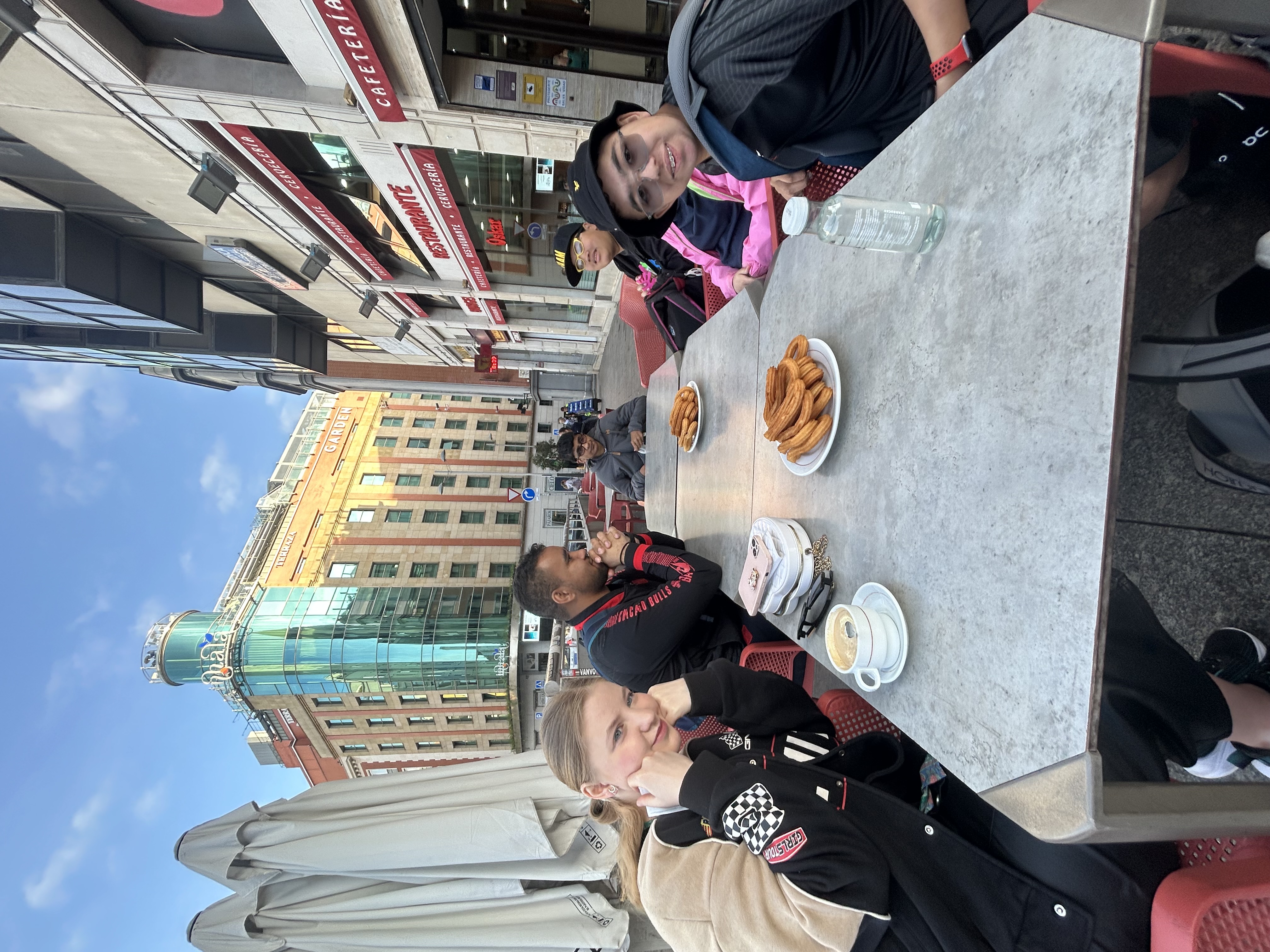 the group sit around tables outside at a pavement cafe. There are coffee cups and plates of churros on the tables