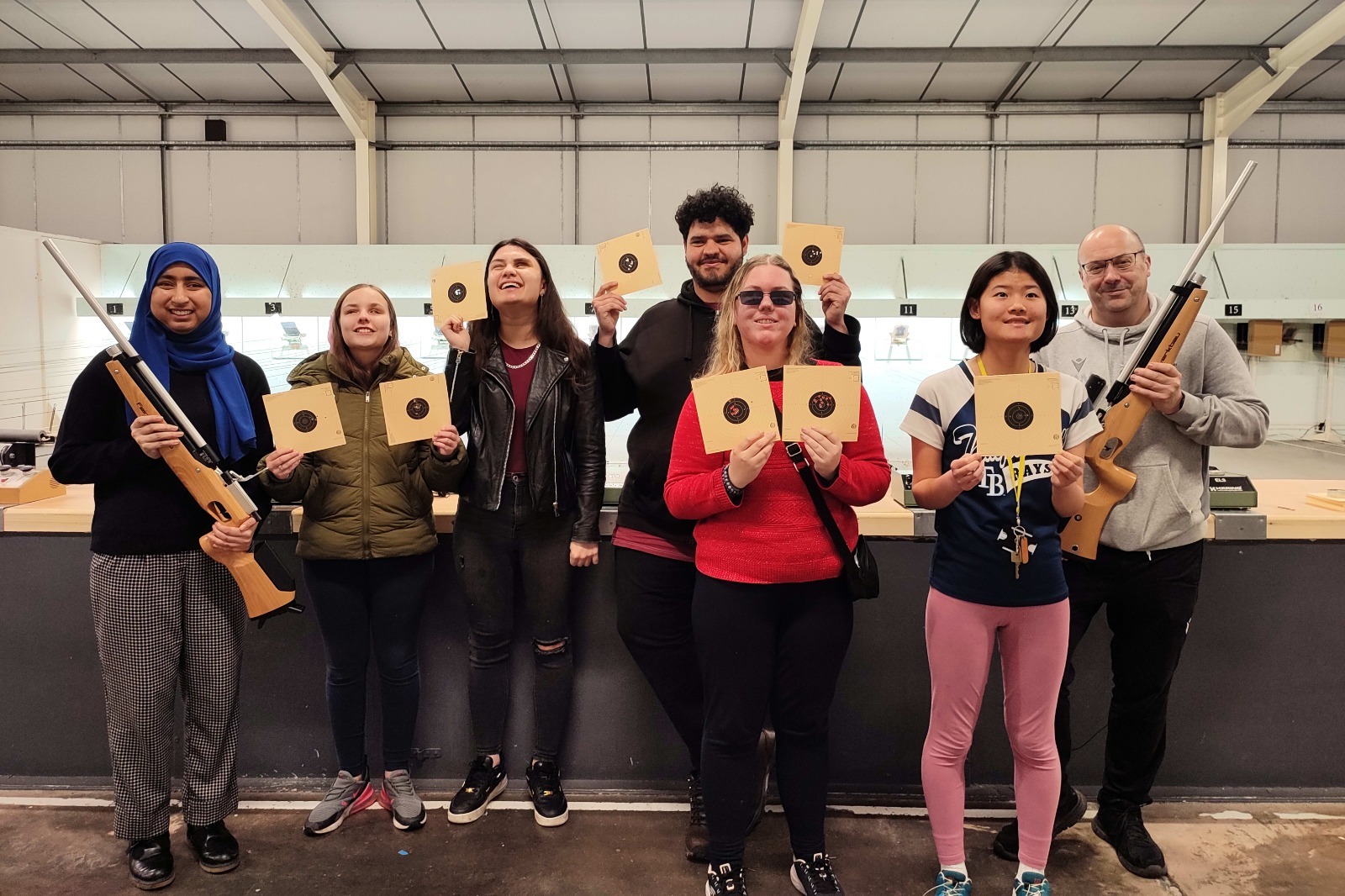 the group of students with coach Jim holding up their targets. On the left a student holds up a rifle and on the right the coach holds up a rifle