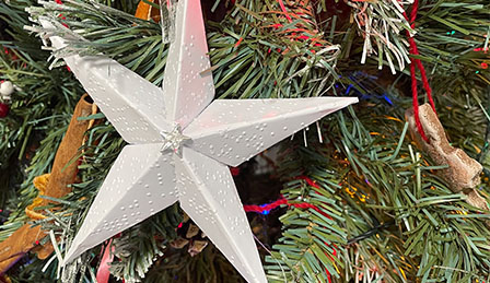 A close up of a star made out of white paper with Braille embossed on it, hanging on the Christmas tree