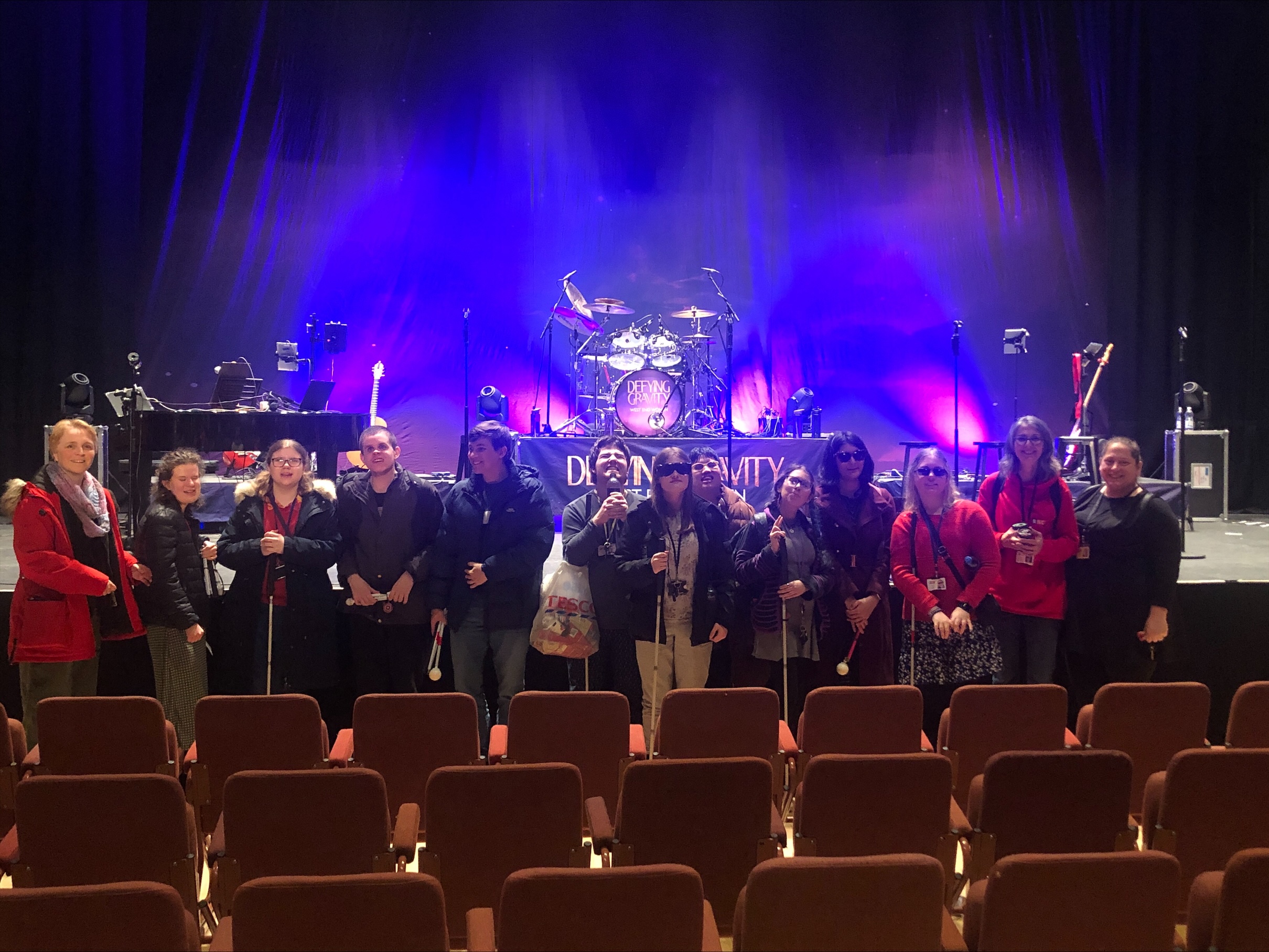 students and volunteers stand in front of an empty stage with words 'Defying Gravity' lit up behind them