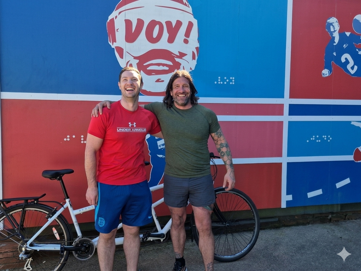 Owen and Gareth with their tandem bike standing in front of student graffiti art which features Owen wearing blind football eyeshades above them
