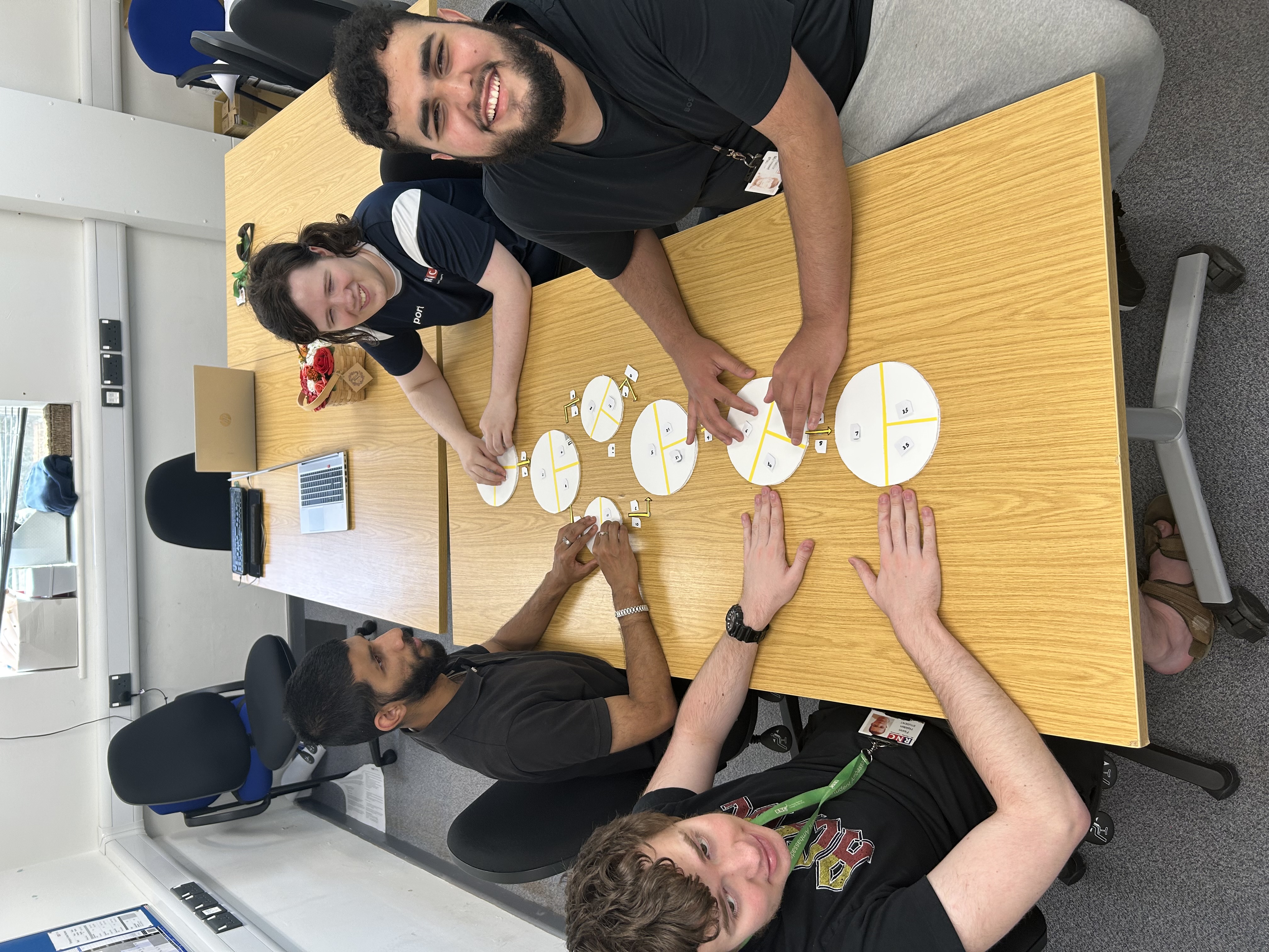 A group of four students sitting around a rectangular wooden table in a classroom setting. They are engaged in an activity involving circular paper pieces divided into sections, for a learning exercise. The students are smiling.