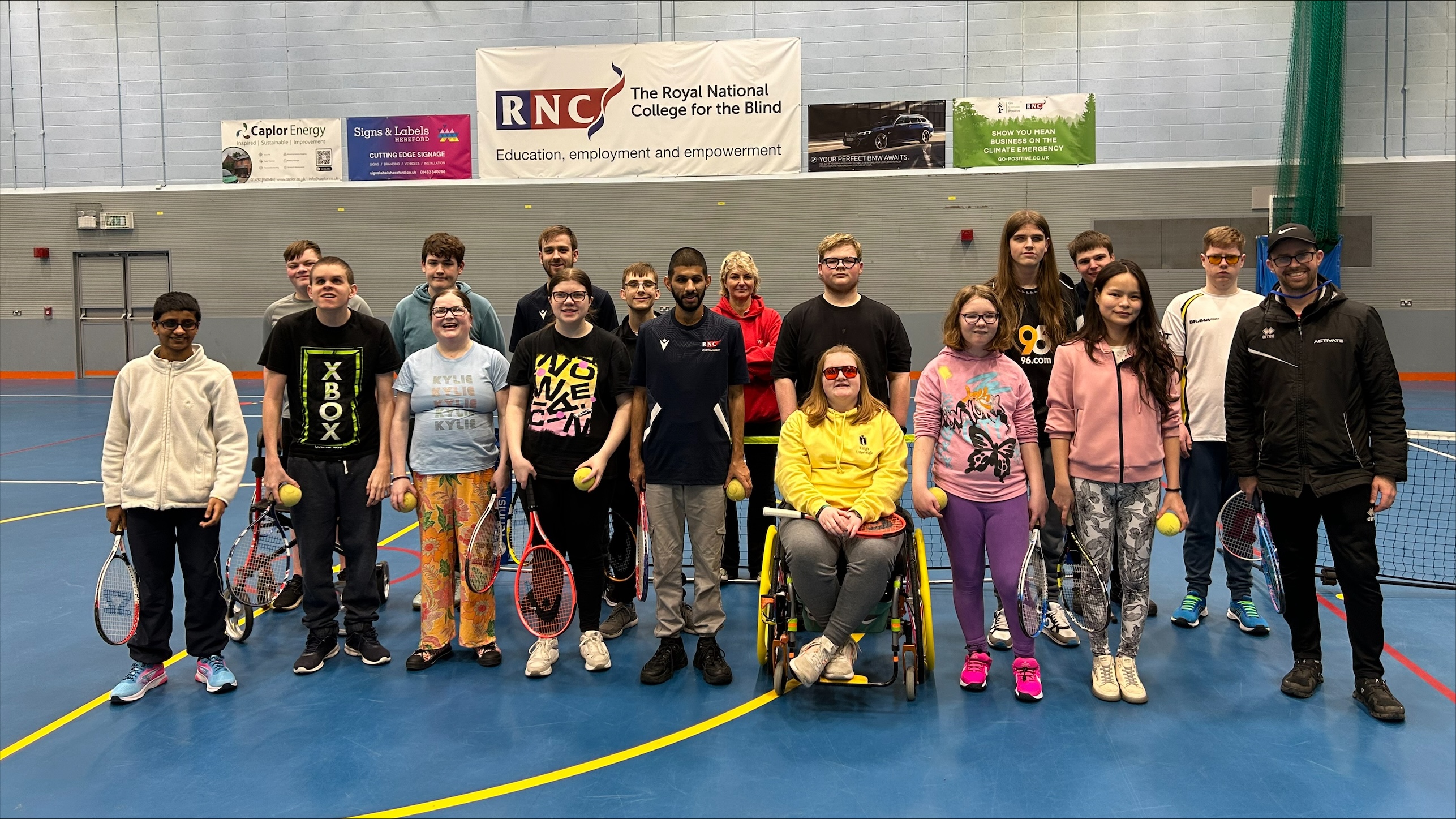 a group of staff, student and tennis coach stand in the sports hall. They all hold tennis racquets and tennis ball 